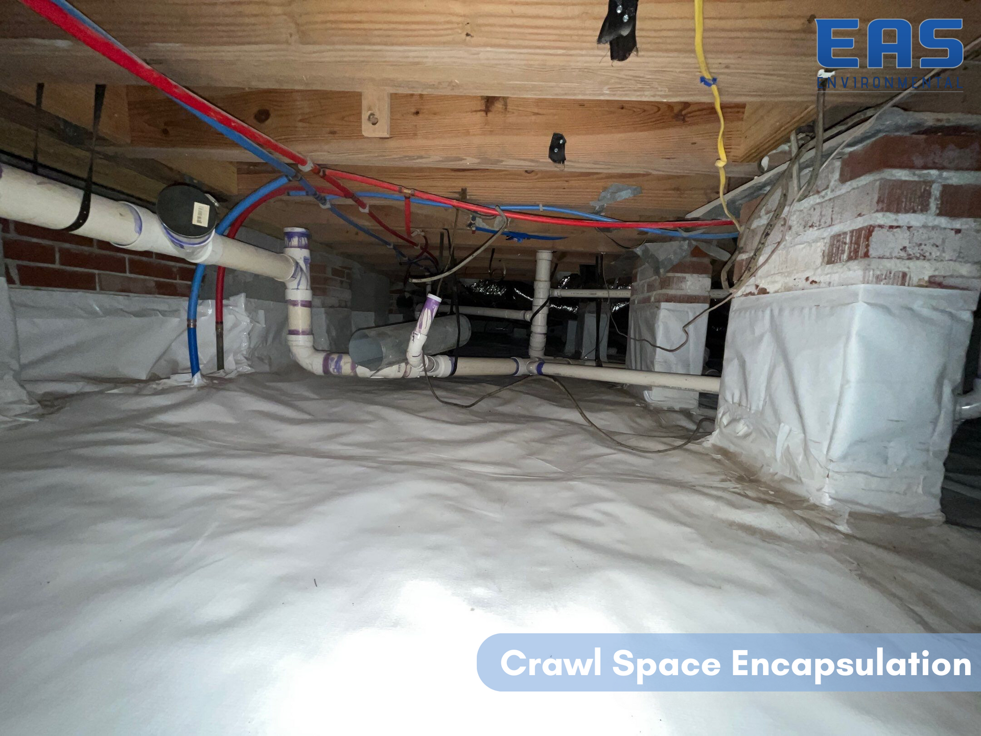 Crawl space with white vapor barrier, pipes, and wiring. Wood beams and brick foundation visible.