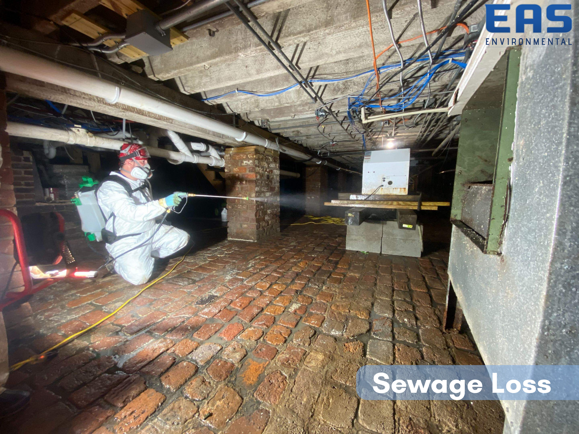 Man in protective suit sprays under a building with sewage loss. Red brick floor, pipes above.