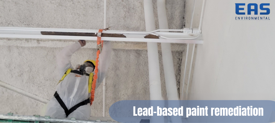 Person in protective suit and respirator removing lead-based paint from a ceiling with white pipes.