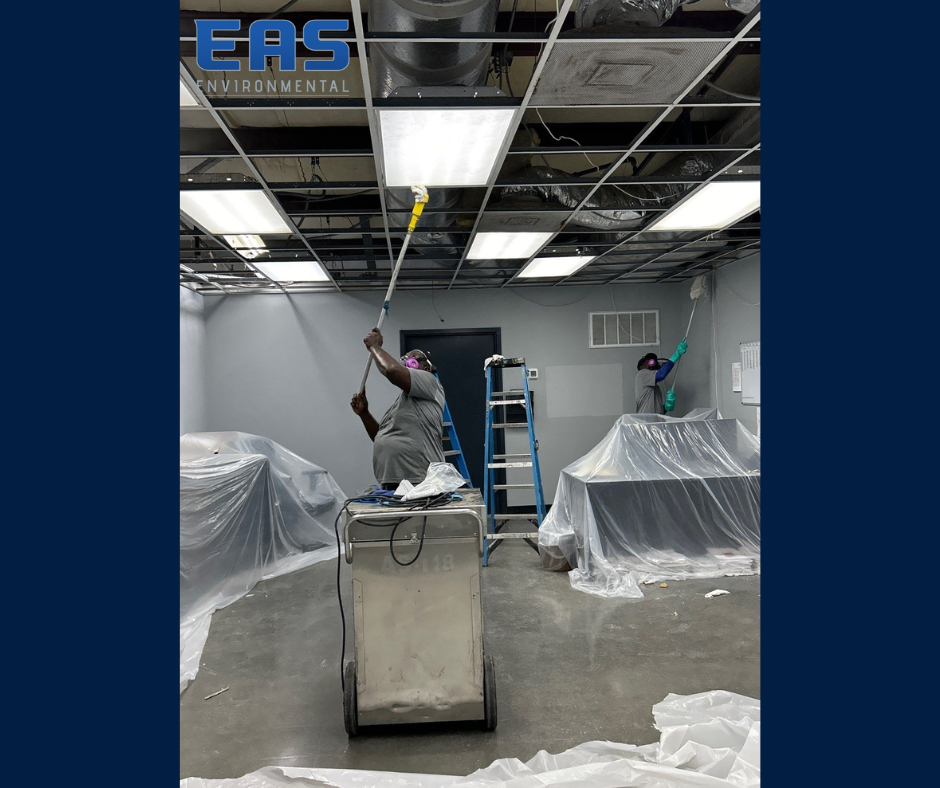 Workers painting a room with drop cloths. One on cart with pole, other on table in room with exposed ceiling.
