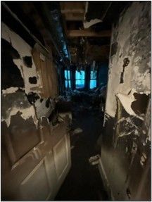 Interior hallway heavily damaged by fire; charred walls, debris, and visible windows at the end.