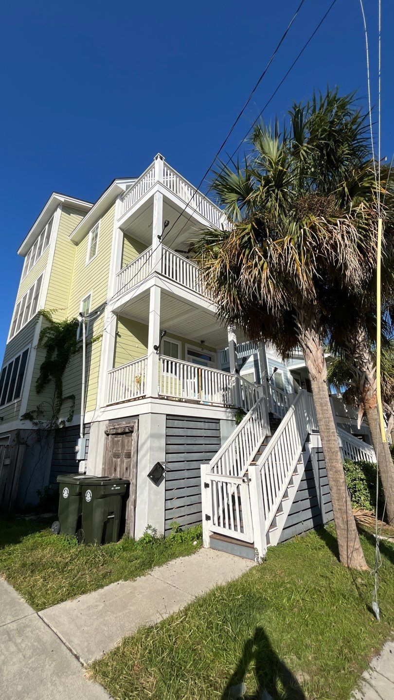 Three-story yellow house with balconies, white trim, and palm trees in front under a blue sky.