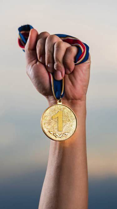 Hand clenching a gold medal with a blue and red ribbon — NQ Plaques & Trophies In Mount Isa, QLD