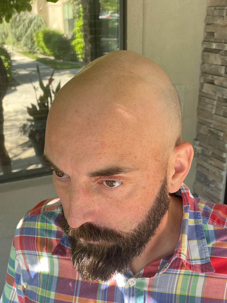 A person with a beard and a shaved head, wearing a colorful plaid shirt, stands outdoors.