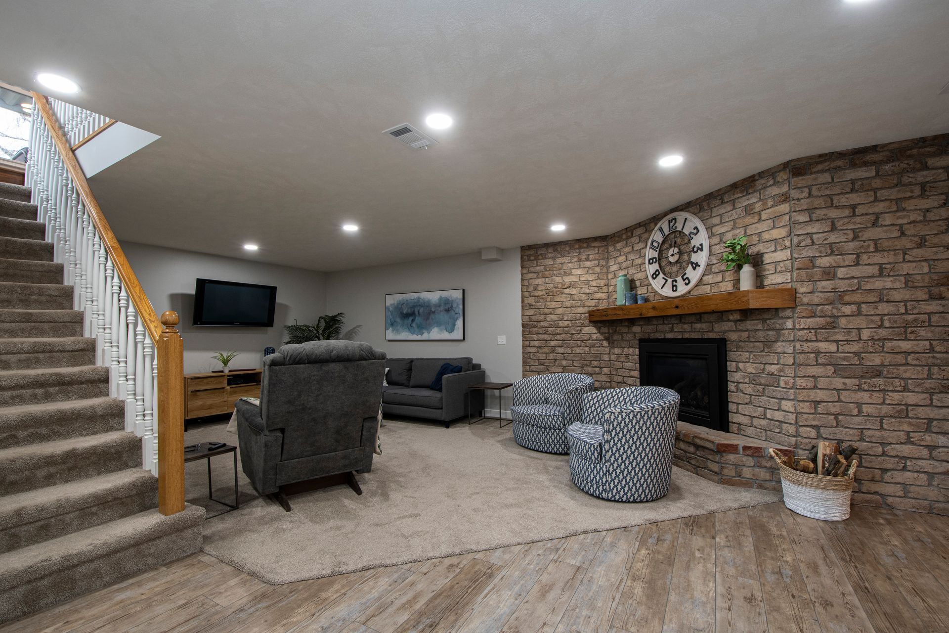 Basement living room with stairs, brick fireplace, recliners, and a desk area