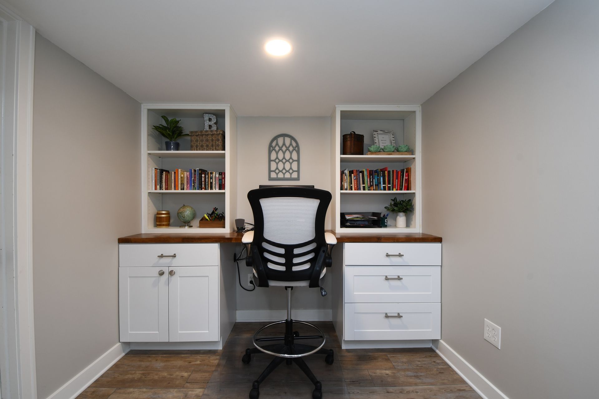Small home office with a black chair, desk, shelves, and white cabinets in a narrow room.