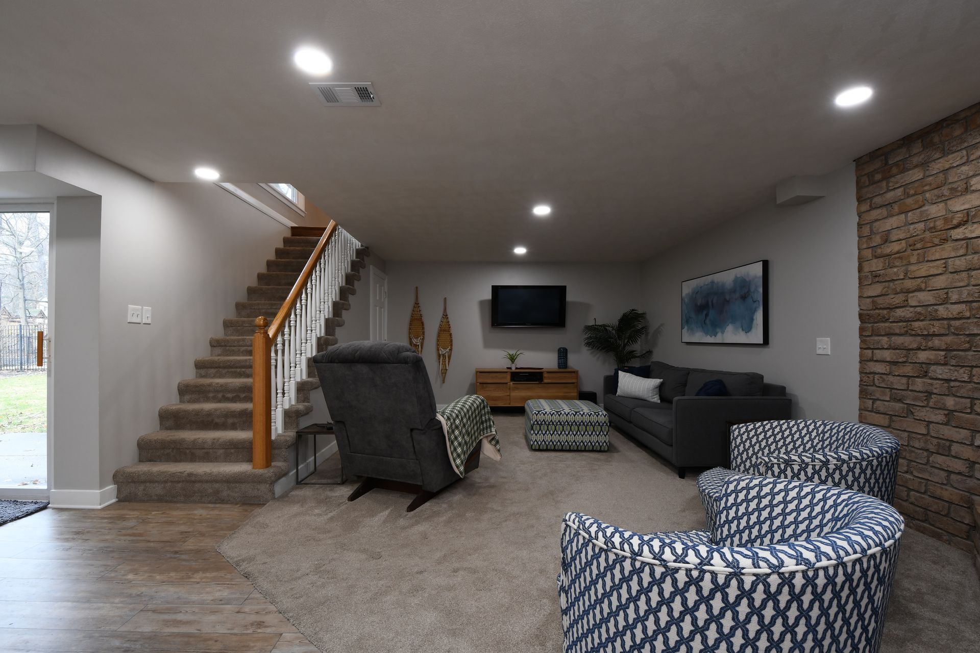 Spacious living room with staircase, dark sofas, patterned chairs, and wall-mounted TV
