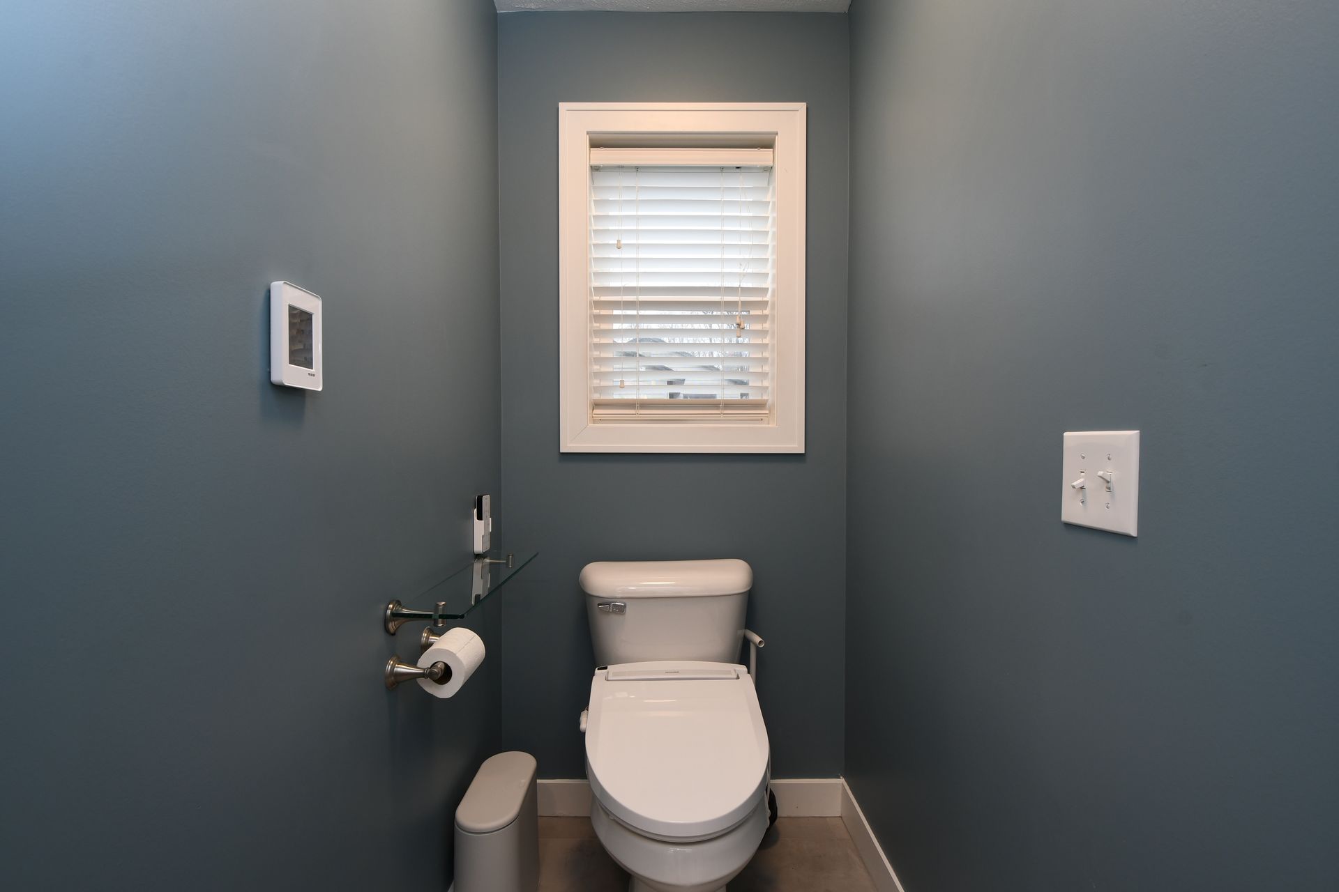 Small blue bathroom with a toilet, toilet paper holder, and window above it.