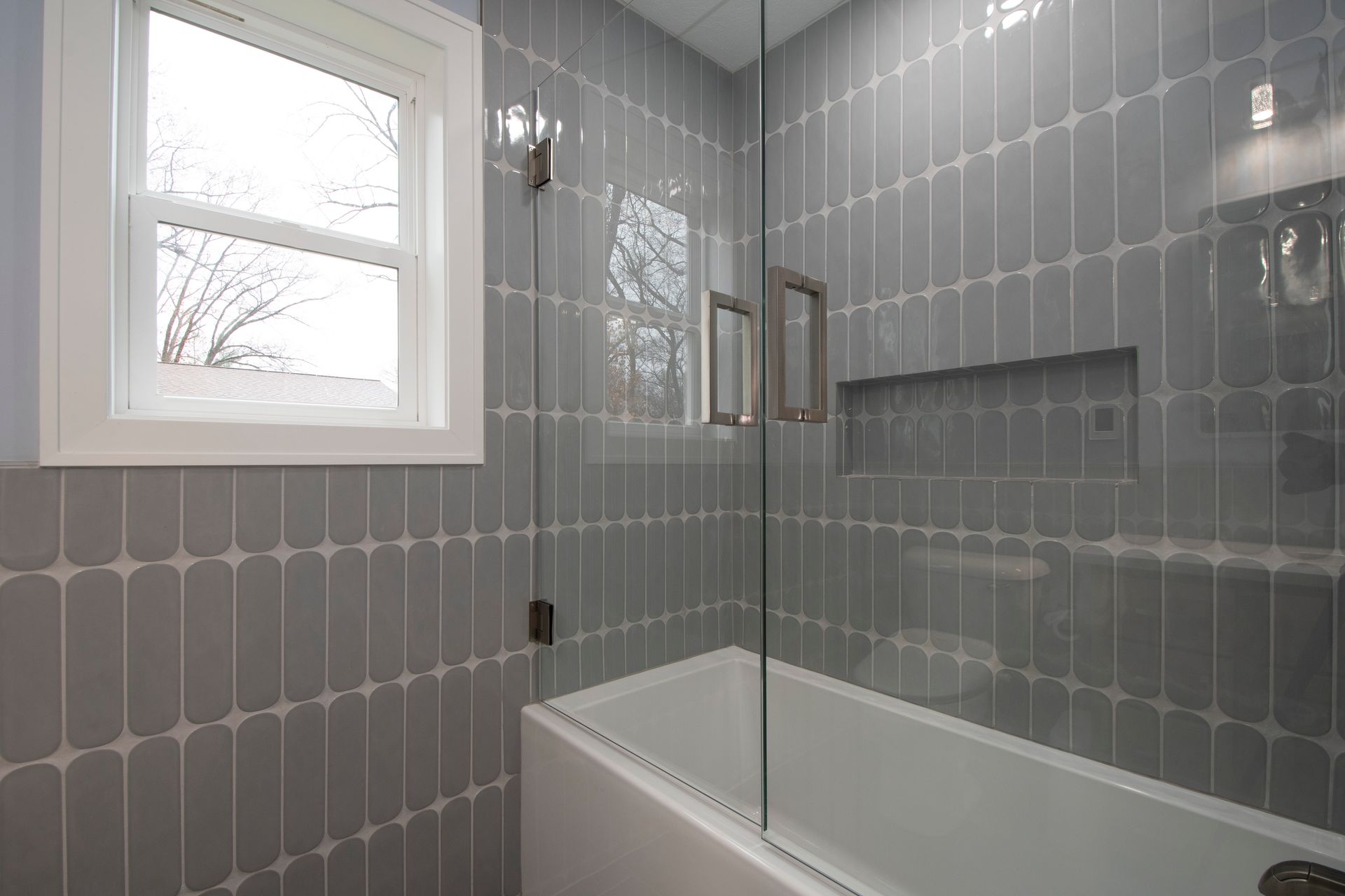 Modern gray-tiled bathroom with a glass shower and white bathtub beside a window
