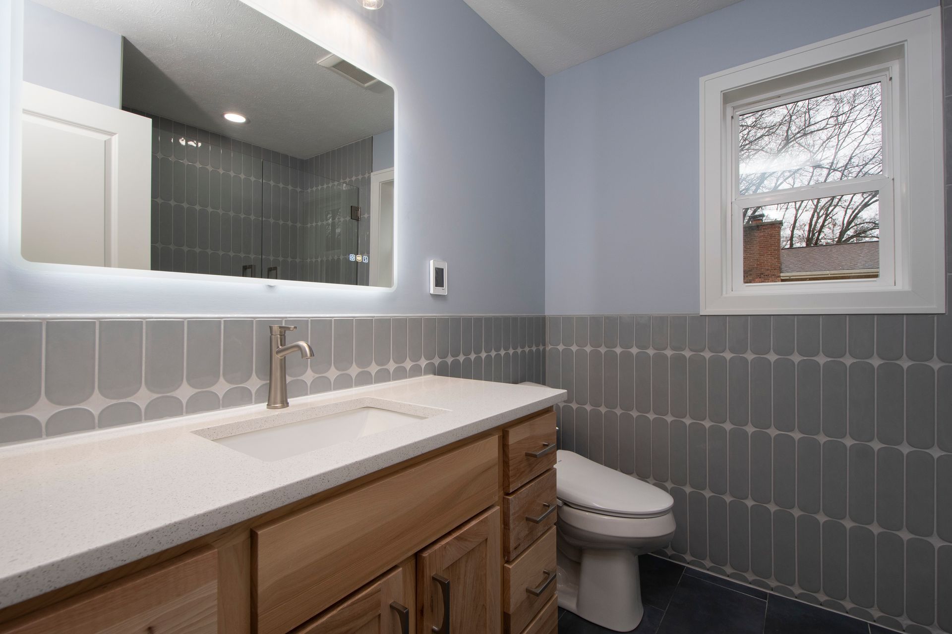 Small bathroom with white vanity, large mirror, gray tile walls, and a window above the toilet.