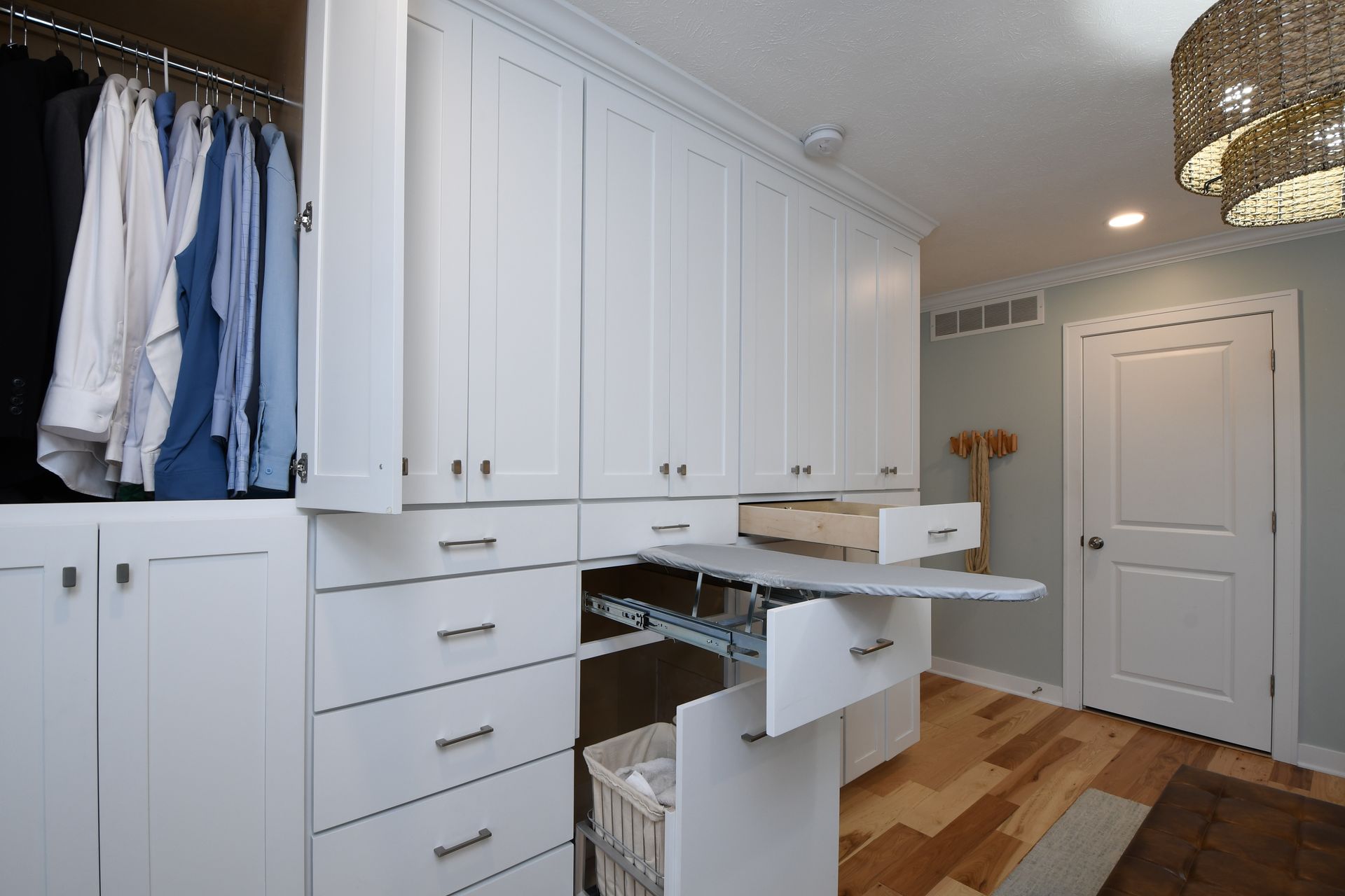 Bright white laundry room with cabinets, hanging clothes, and a pull-out ironing board on a wood floor.