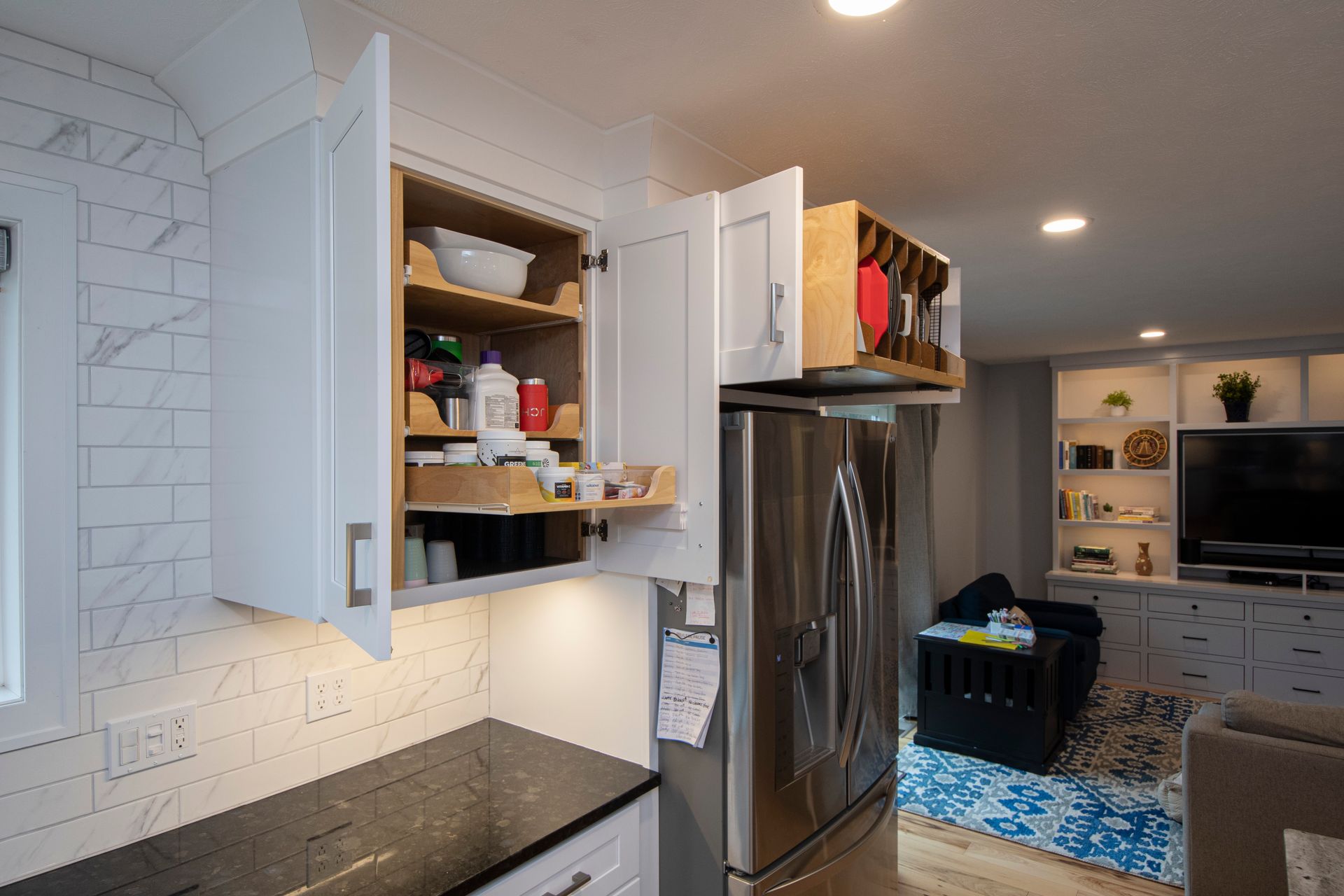 Kitchen with open white cabinets, stainless fridge, and view into a living room with blue rug and shelves