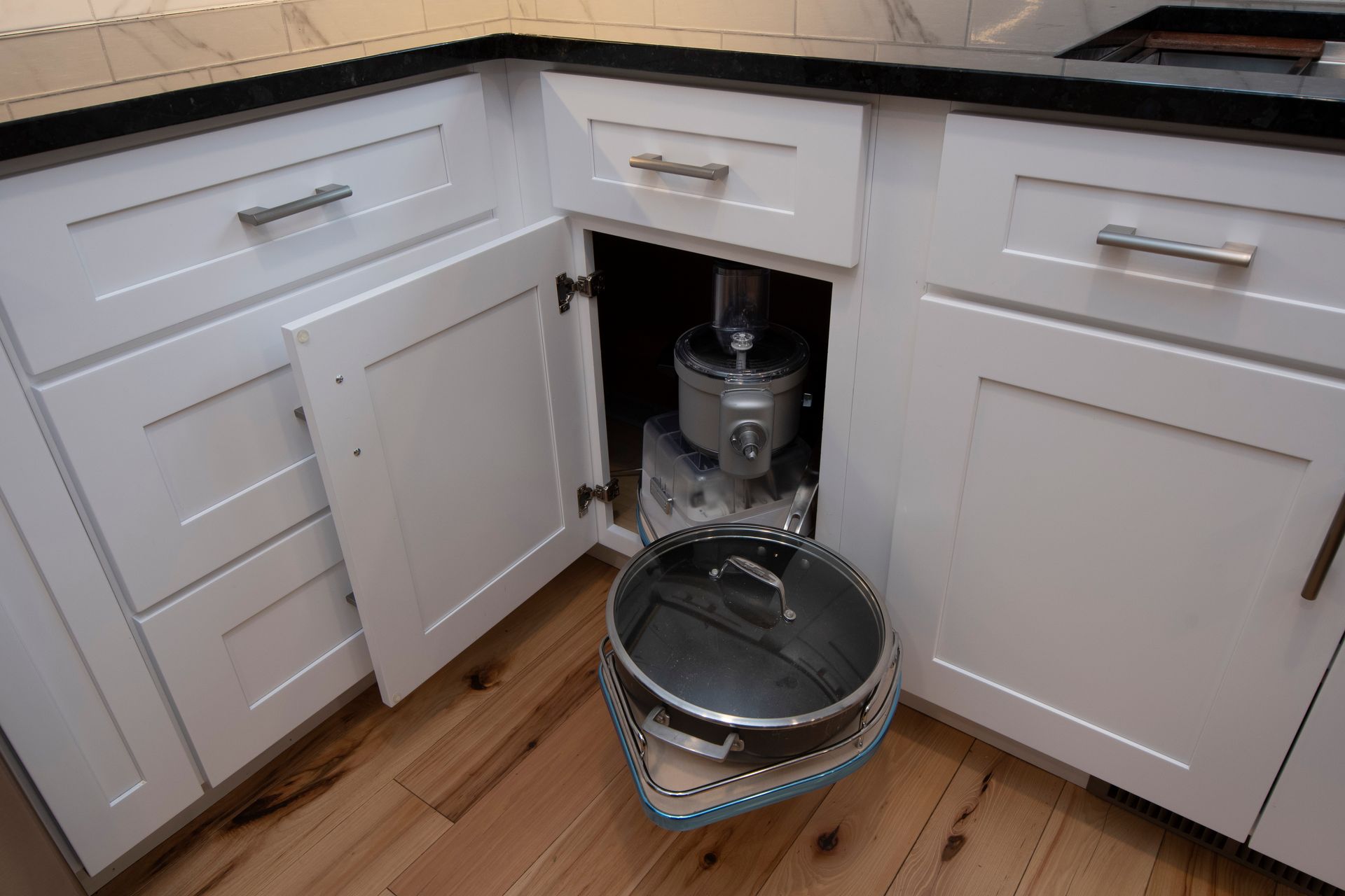 Kitchen cabinet under sink open, revealing plumbing and a metal bucket on the floor.