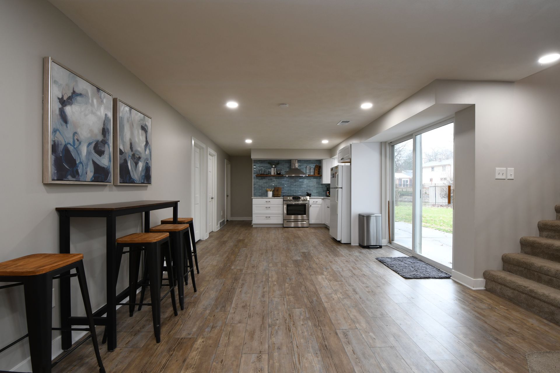 Modern open-plan living area with bar stools, kitchen, stairs, and sliding glass door to outside.