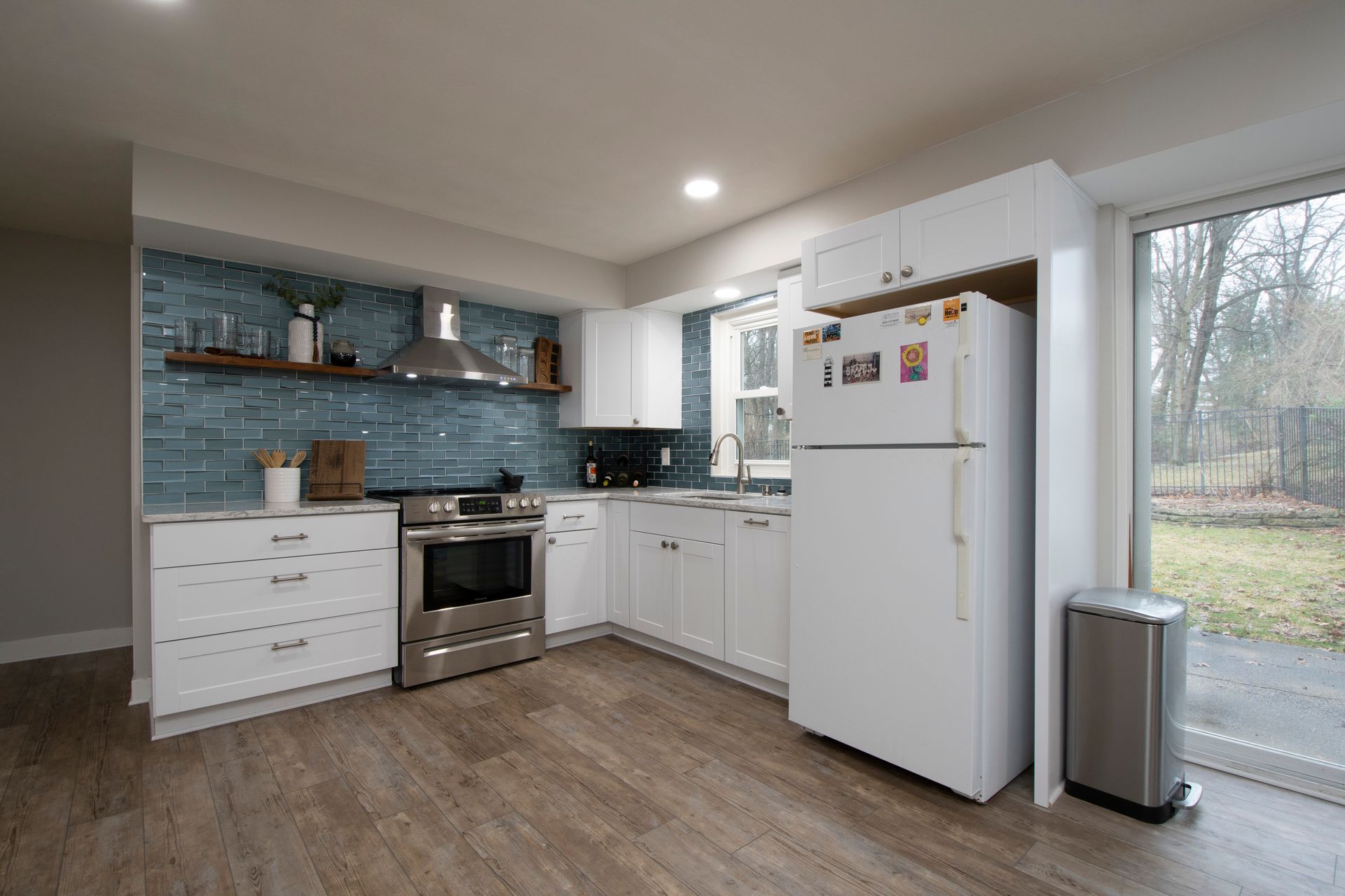 Modern white kitchen with blue tile backsplash, stainless oven, white fridge, and sliding glass door.