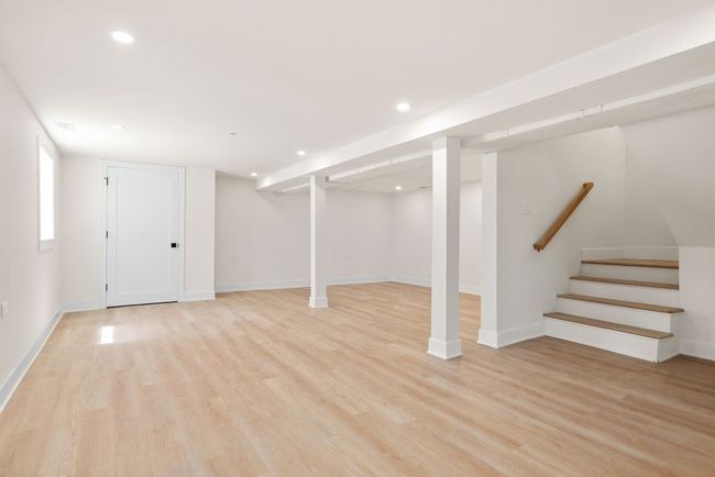Bright, empty basement room with light wood floors, white walls, recessed lights, and a staircase.