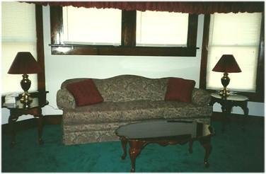 Living room with patterned sofa, two lamps, windows, and a coffee table on green carpet