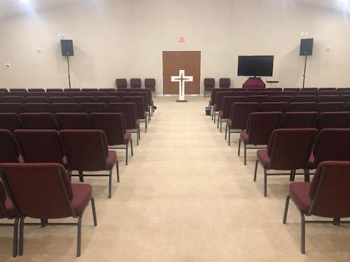 Empty church sanctuary with rows of maroon chairs facing a white cross and pulpit.