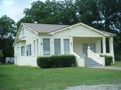 Small white house with gray shutters, front steps, and a green lawn under trees