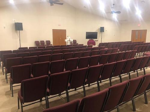 Empty auditorium with rows of maroon chairs, beige walls, stage speakers, and a wooden door at the front.