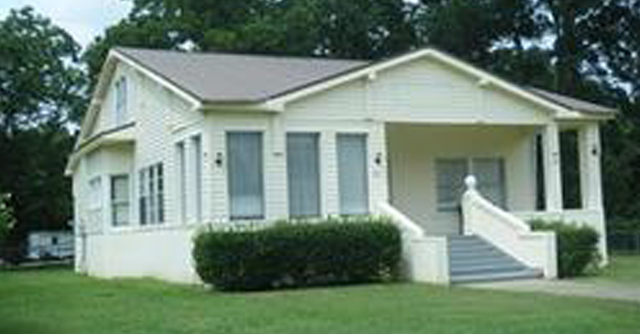 Small white house with a front porch, steps, and trimmed bushes on a green lawn