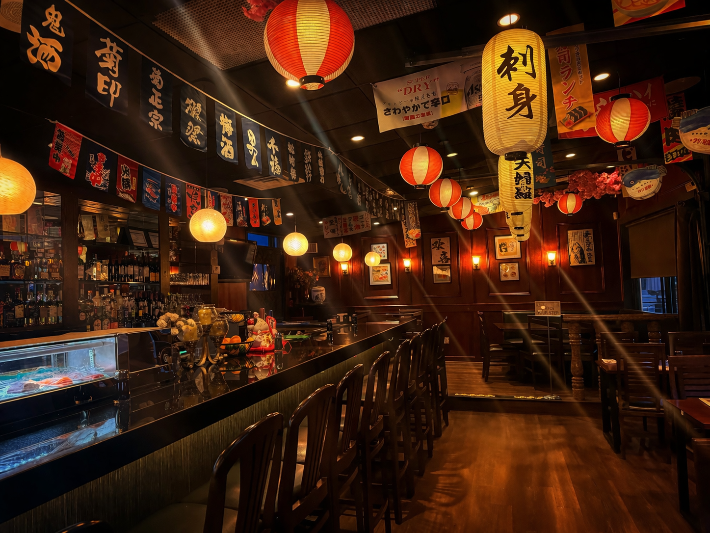 An indoor view of a dimly lit Japanese restaurant, featuring a long counter, bar stools, and hanging paper lanterns.