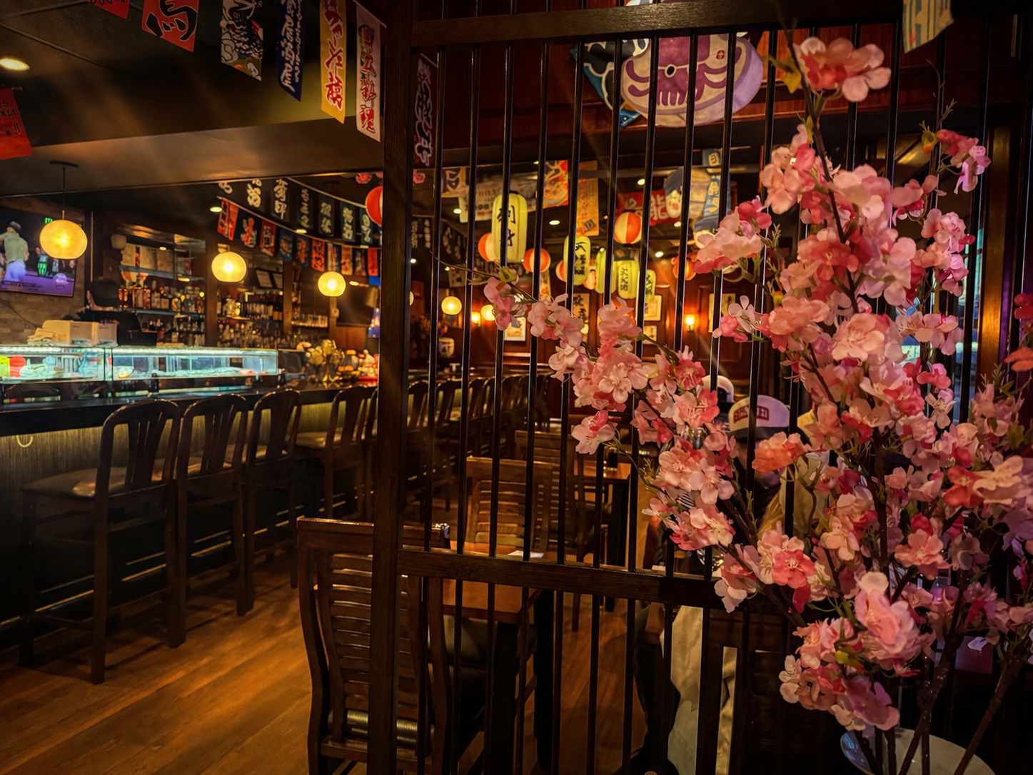 An interior view of a Japanese-style restaurant with a bar, wooden furniture, hanging banners, and faux cherry blossoms.