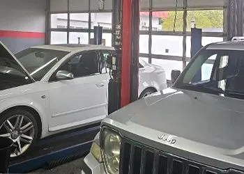 White sedan and silver Jeep parked inside an auto shop garage with lifts.