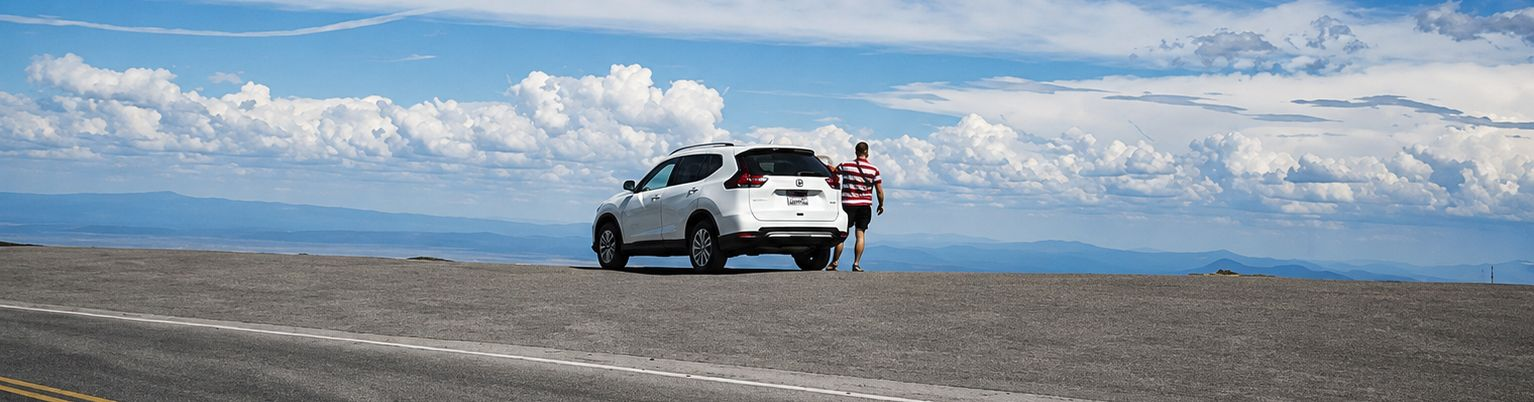 White SUV parked on a rocky shore with two people standing beside it under a cloudy blue sky.