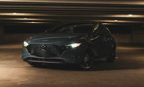 Dark gray Mazda 3 hatchback parked in an underground parking garage with headlights on.