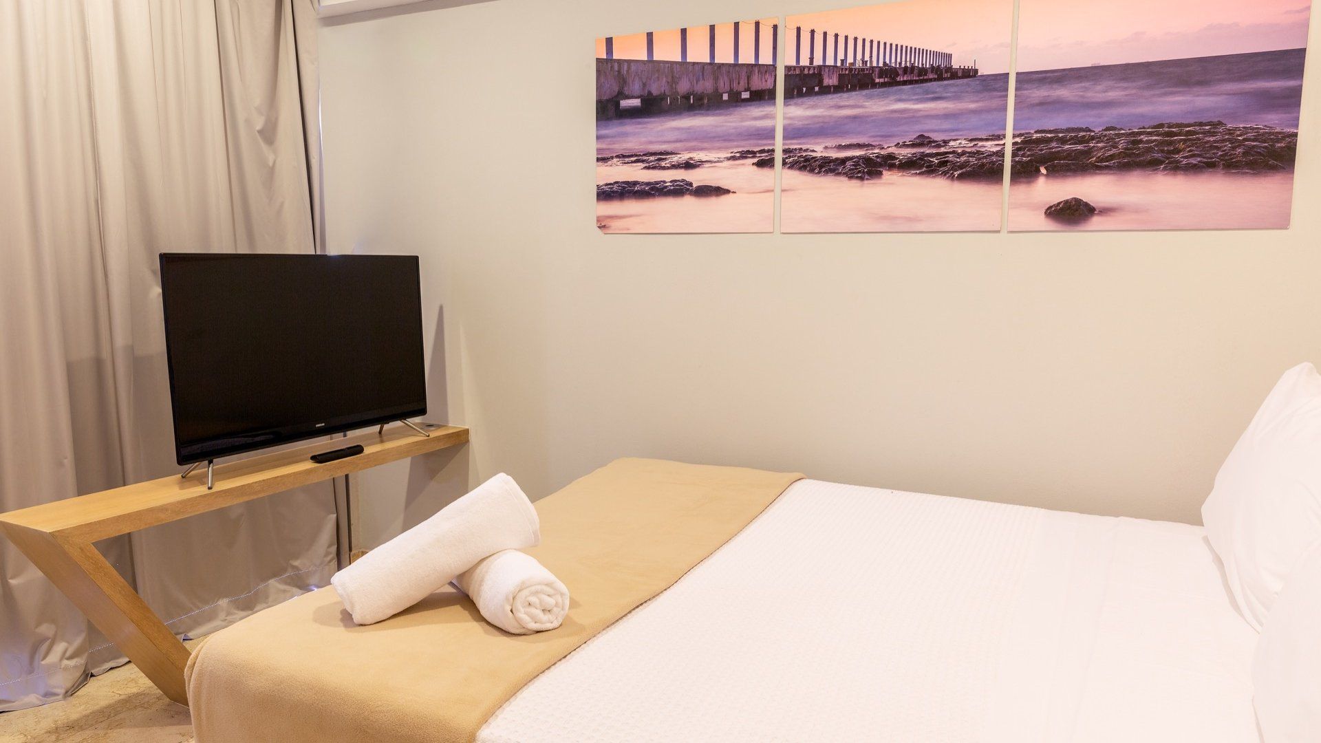 Habitación de hotel con una cama bien hecha, cuadros en la pared, televisor y una toalla doblada sobre un banco.