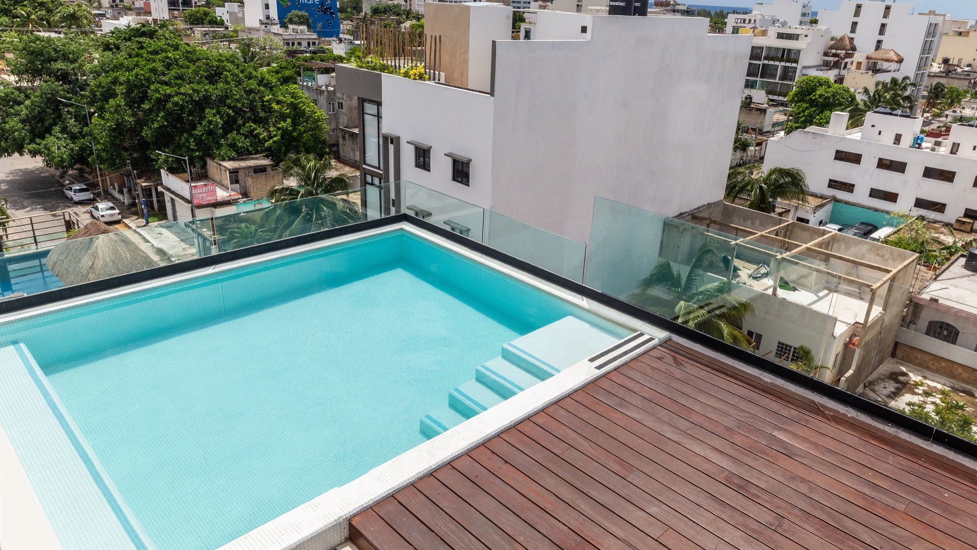 Piscina en la azotea con agua turquesa junto a una terraza de madera y edificios de la ciudad al fondo.