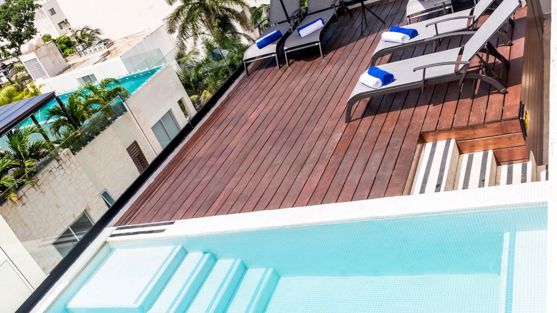 Piscina en la azotea junto a una terraza de madera con tumbonas y vistas al edificio de tonalidad azulada.
