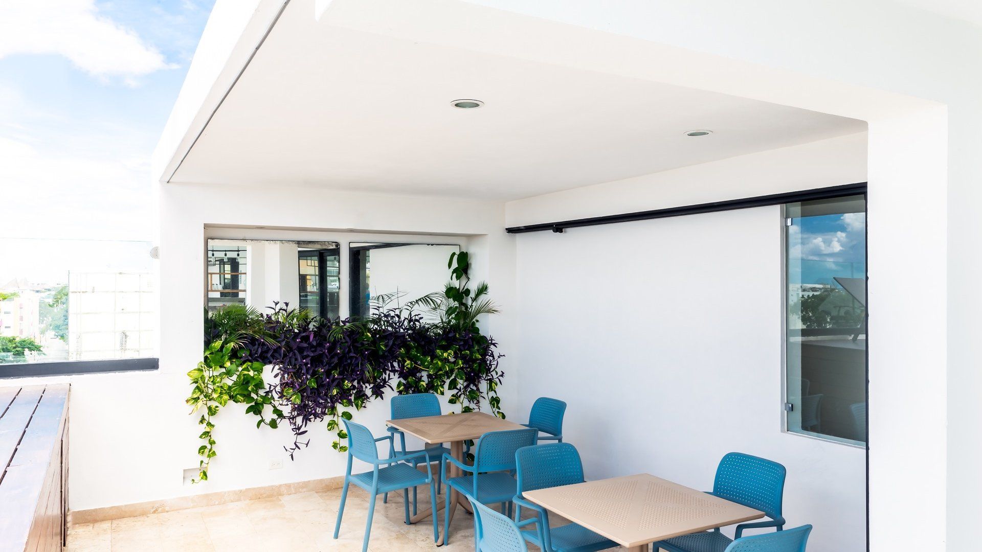 Patio exterior con sillas y mesas azules junto a una pared blanca y flores moradas colgantes.