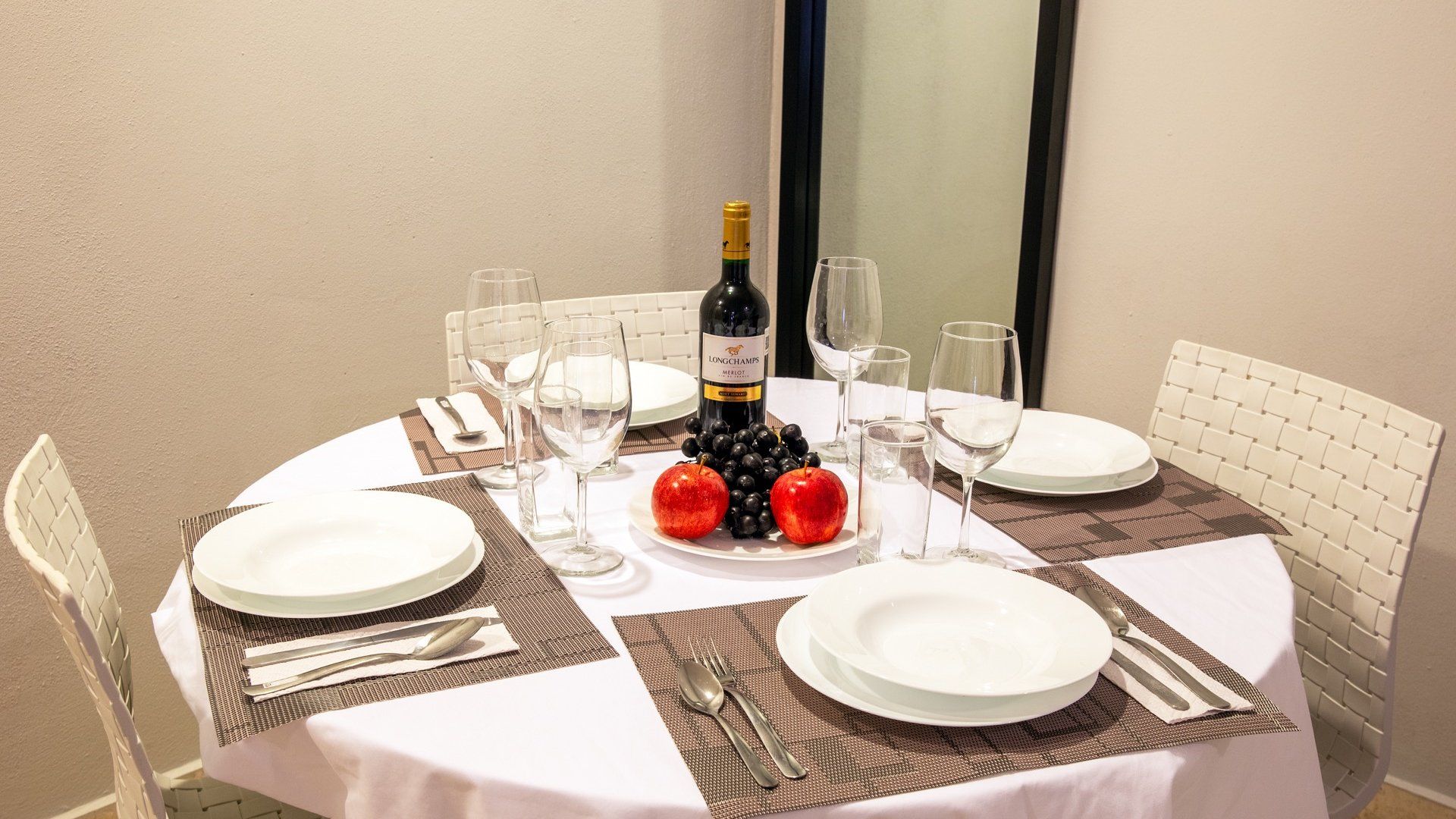 Prepara la mesa del comedor con una botella de vino, copas, platos y velas rojas sobre un mantel blanco.