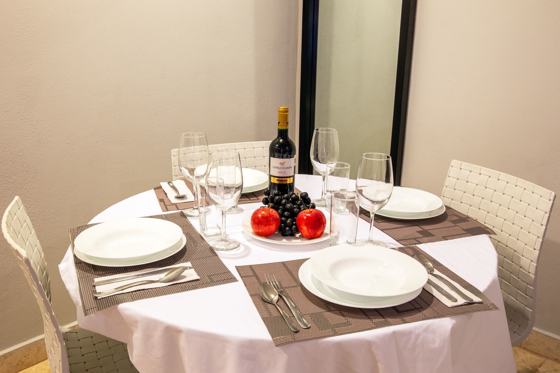 Mesa del comedor con platos blancos, copas de vino, velas y un centro de mesa con frutas en una habitación de la esquina.