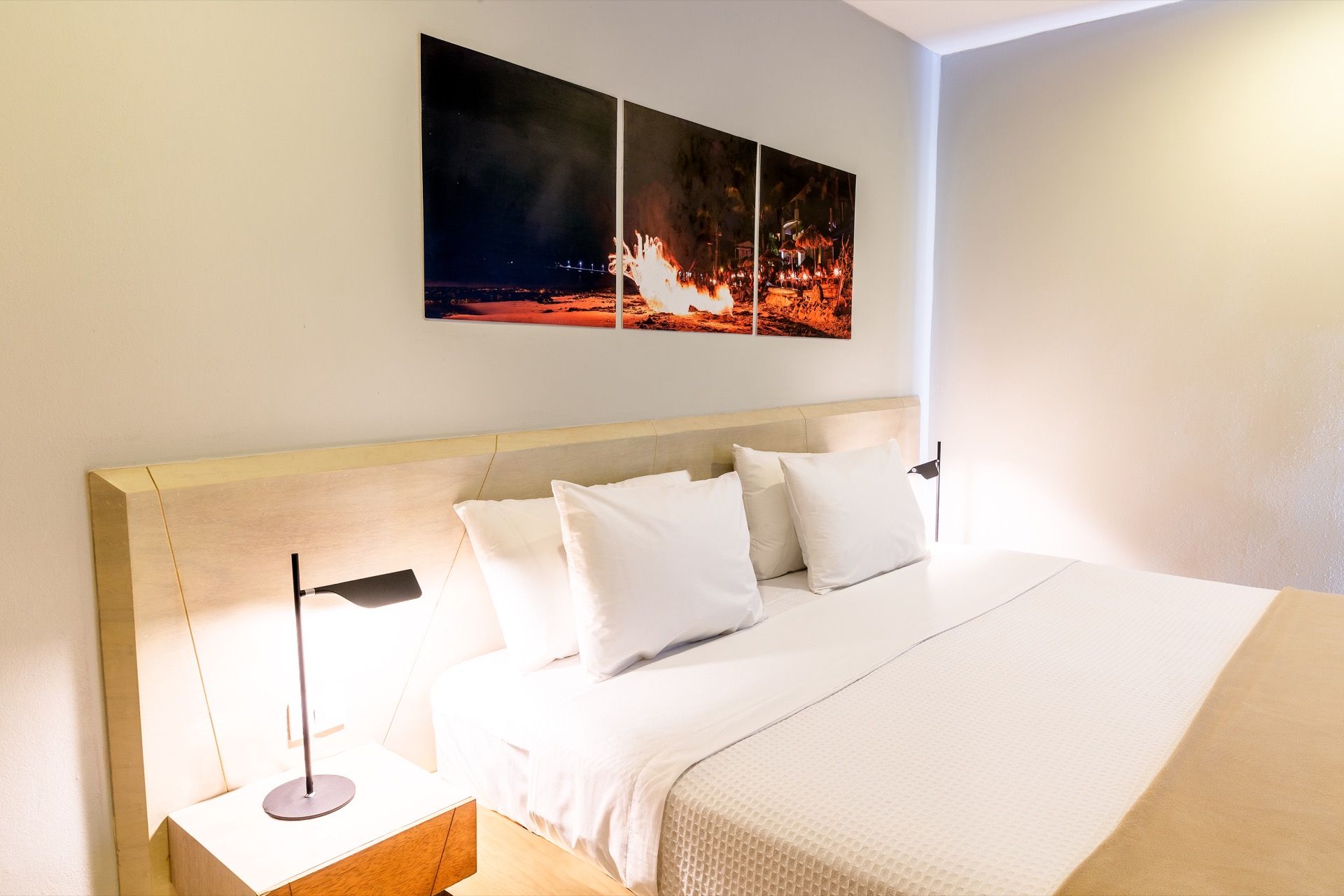 Dormitorio moderno y luminoso con cama blanca, lámpara de noche y cuadro con paisaje urbano.