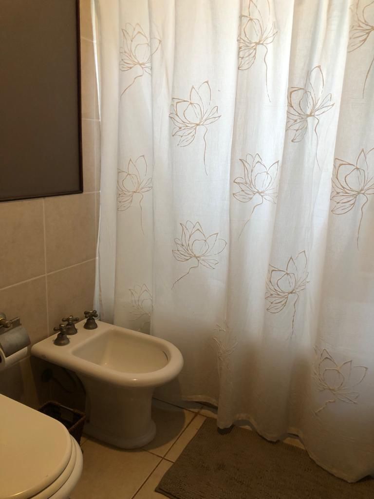 Baño con inodoro blanco, bidé y cortina de ducha con estampado floral. También se aprecian