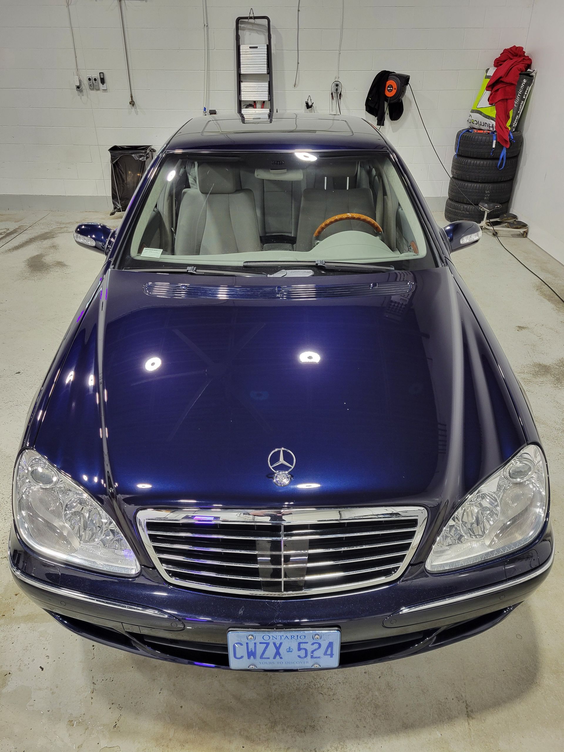 Front view of a dark blue Mercedes sedan parked in a garage, with wall tools and a ladder behind it.