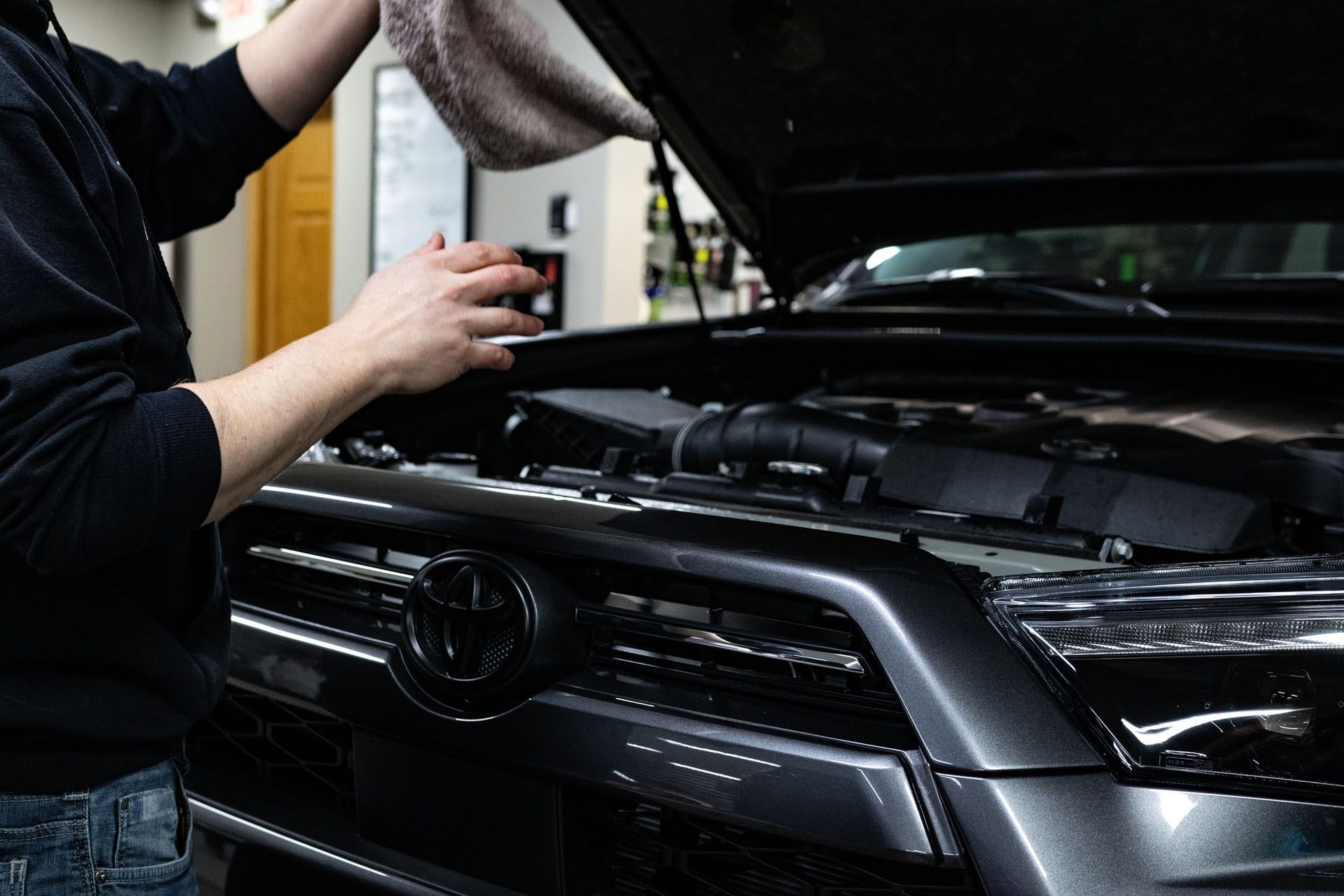 Mechanic working under an open car hood in a garage, inspecting a black vehicle engine bay
