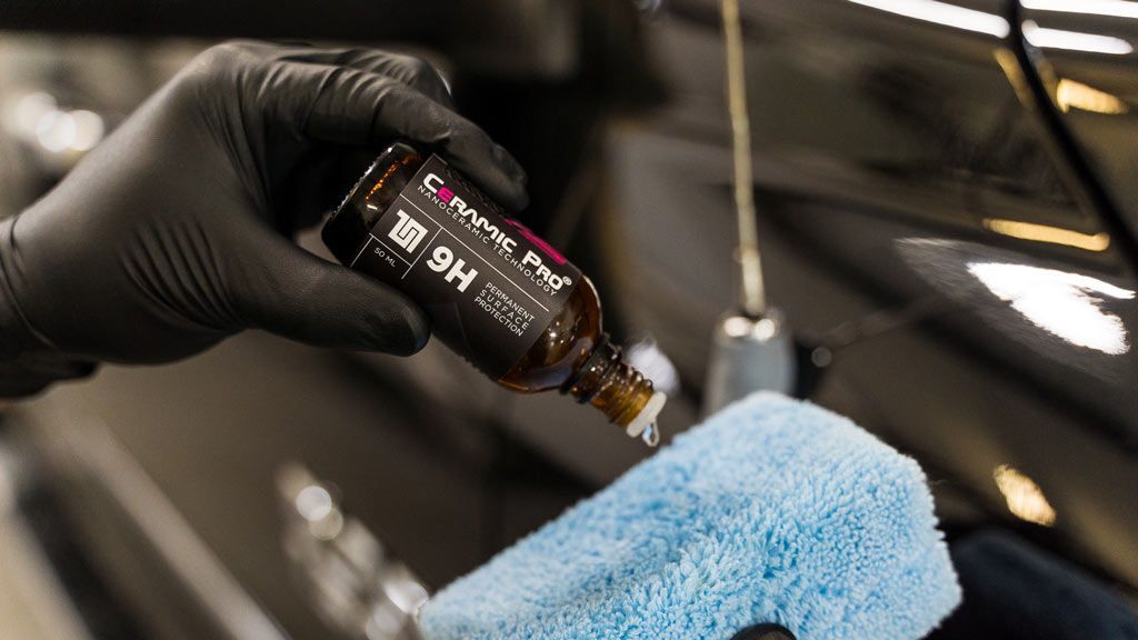 Gloved hand applying car polish with a blue microfiber cloth on a black vehicle.