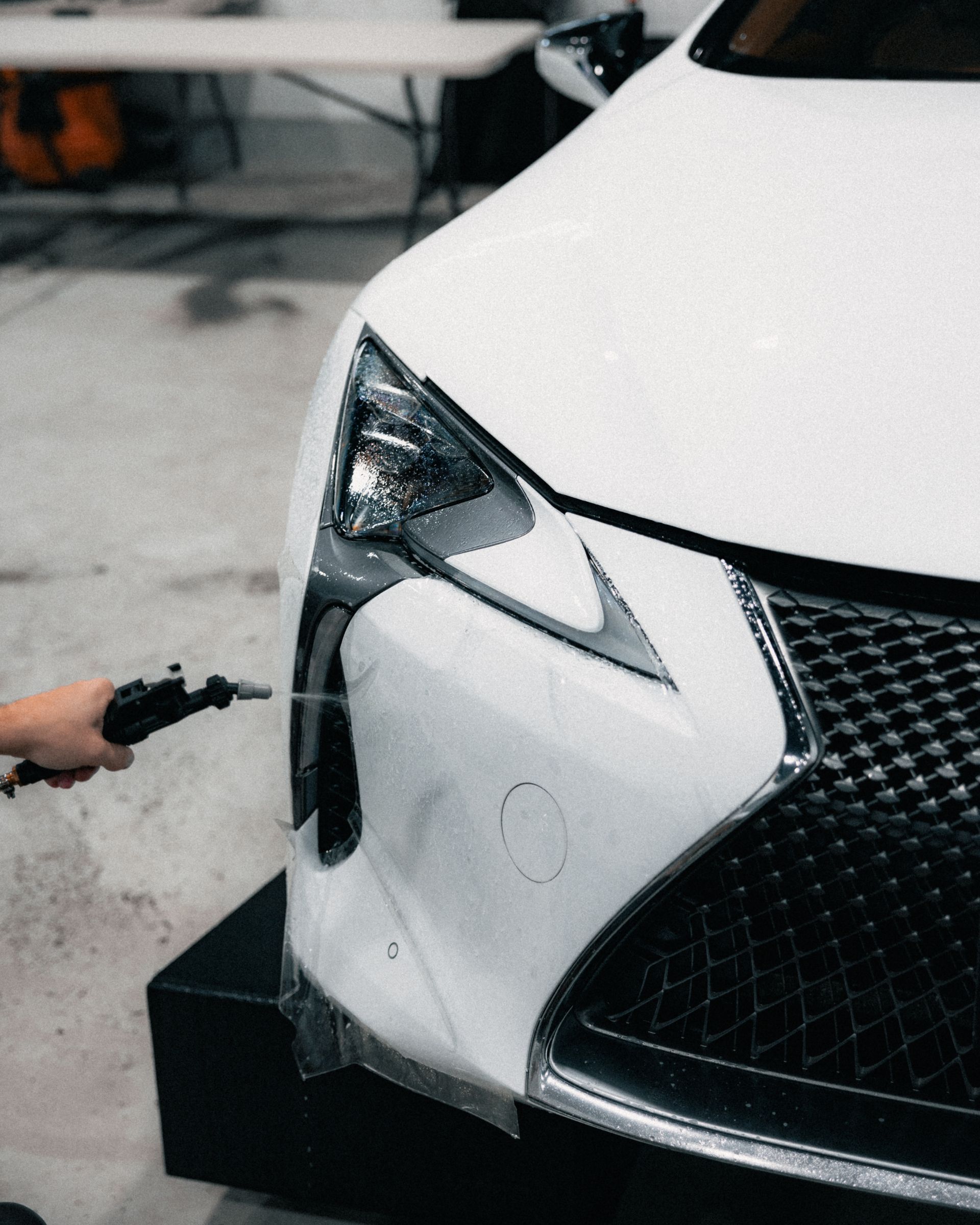 Person holding a spray gun near the front of a white car, likely painting or detailing it