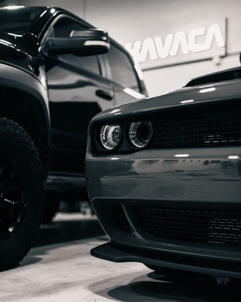 Two black sports cars in a showroom, with a close-up of a gray car’s front grille and headlights.