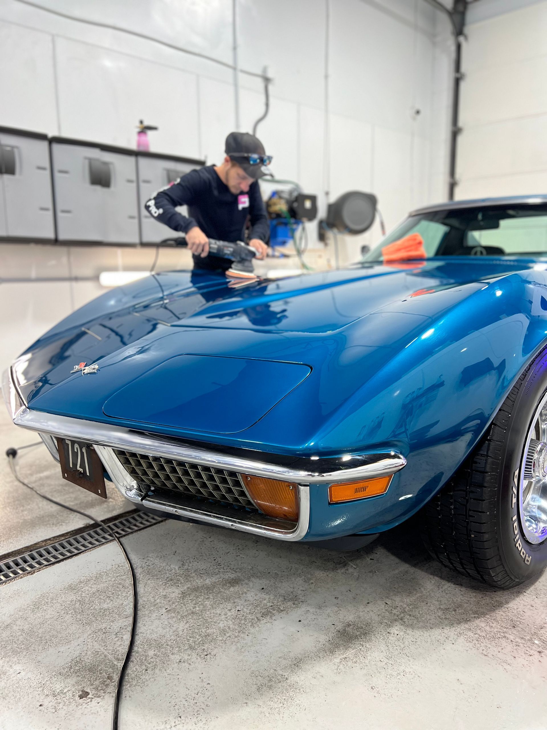 Blue classic car being polished by a worker in a garage, with water on the floor.