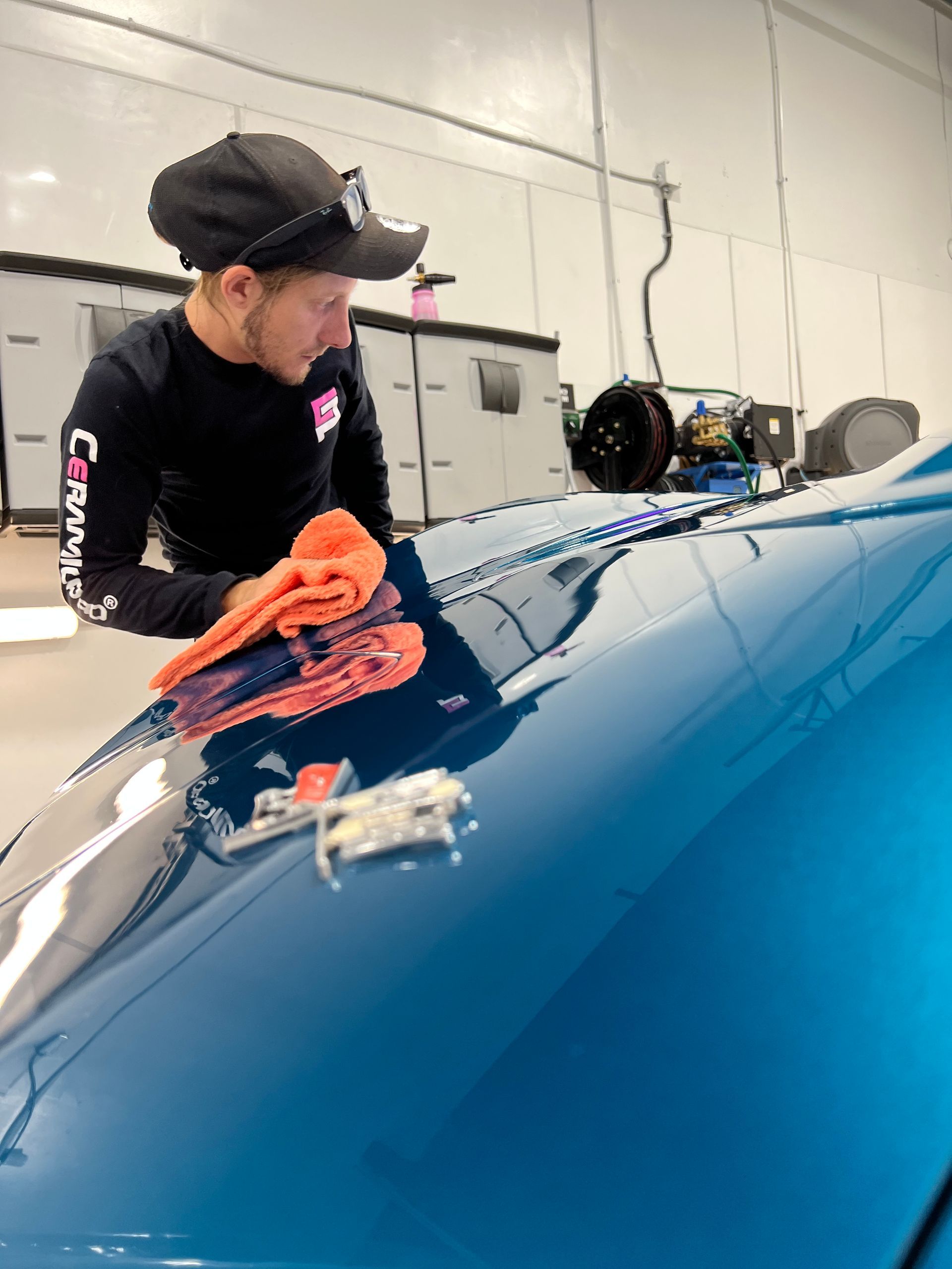 Person polishing a blue car hood with an orange cloth in a garage.