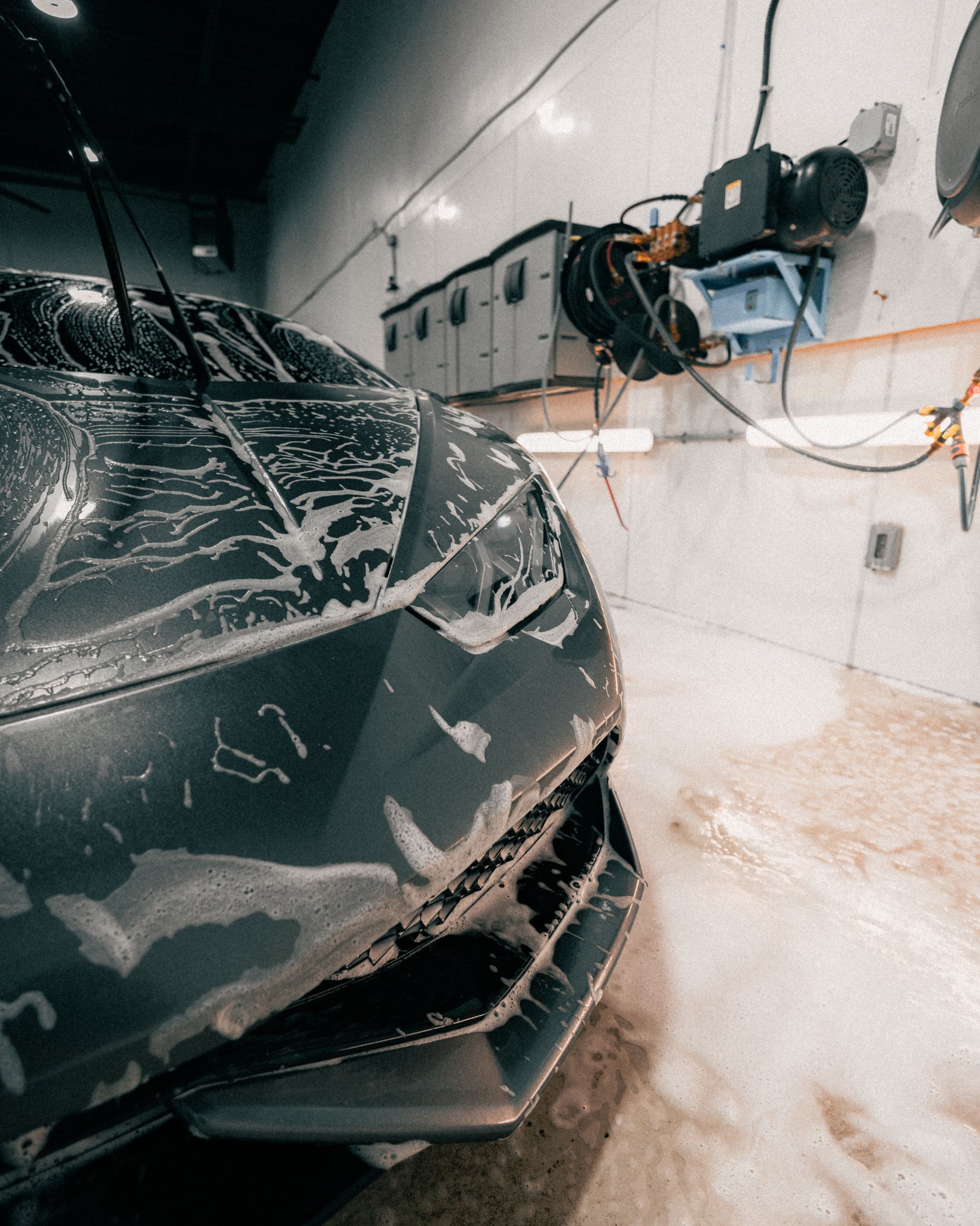 Front of a black car in a garage, covered in soap suds and water spray, near wall-mounted equipment