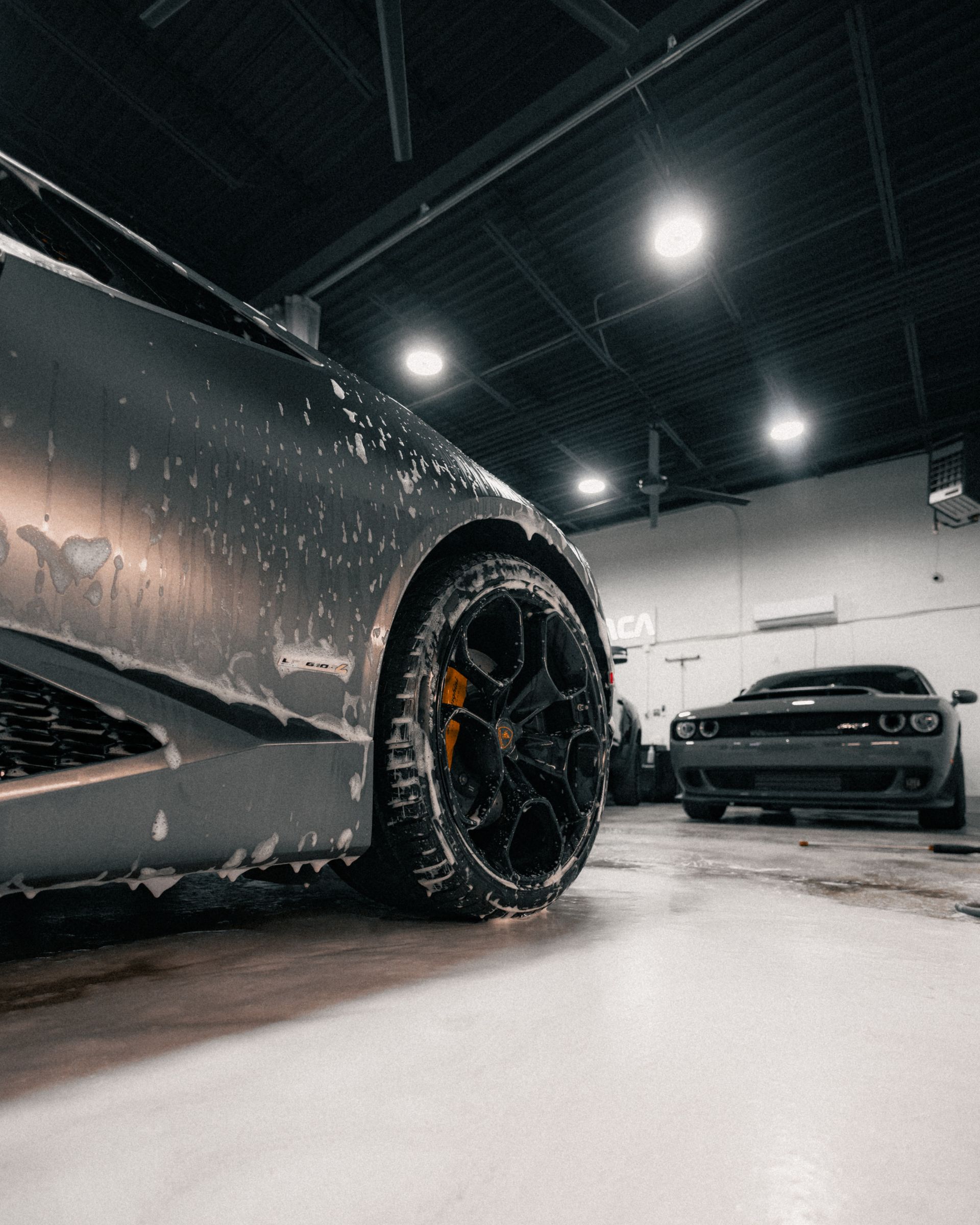 Black sports car in a wet garage, low-angle view of rear wheel and front car in the background