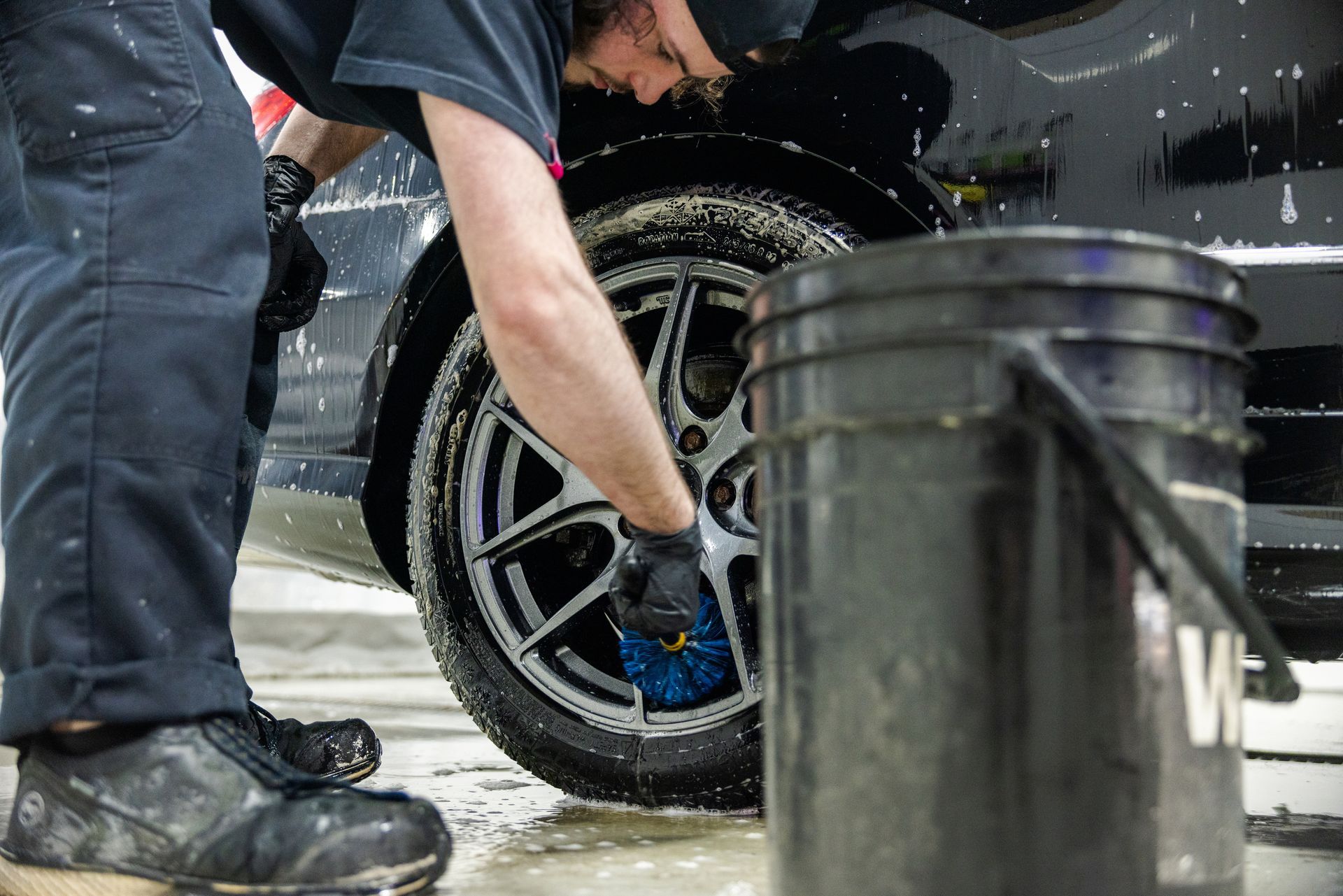 Person cleaning a black car wheel with a blue brush beside a water bucket.