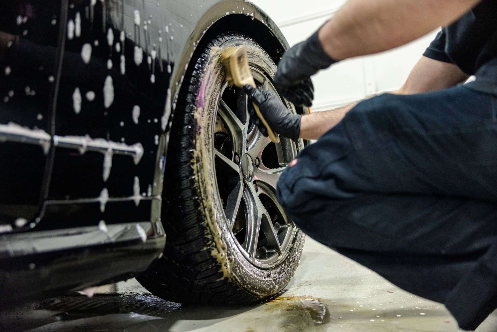 Person washing a black car wheel with soap and water in a garage.