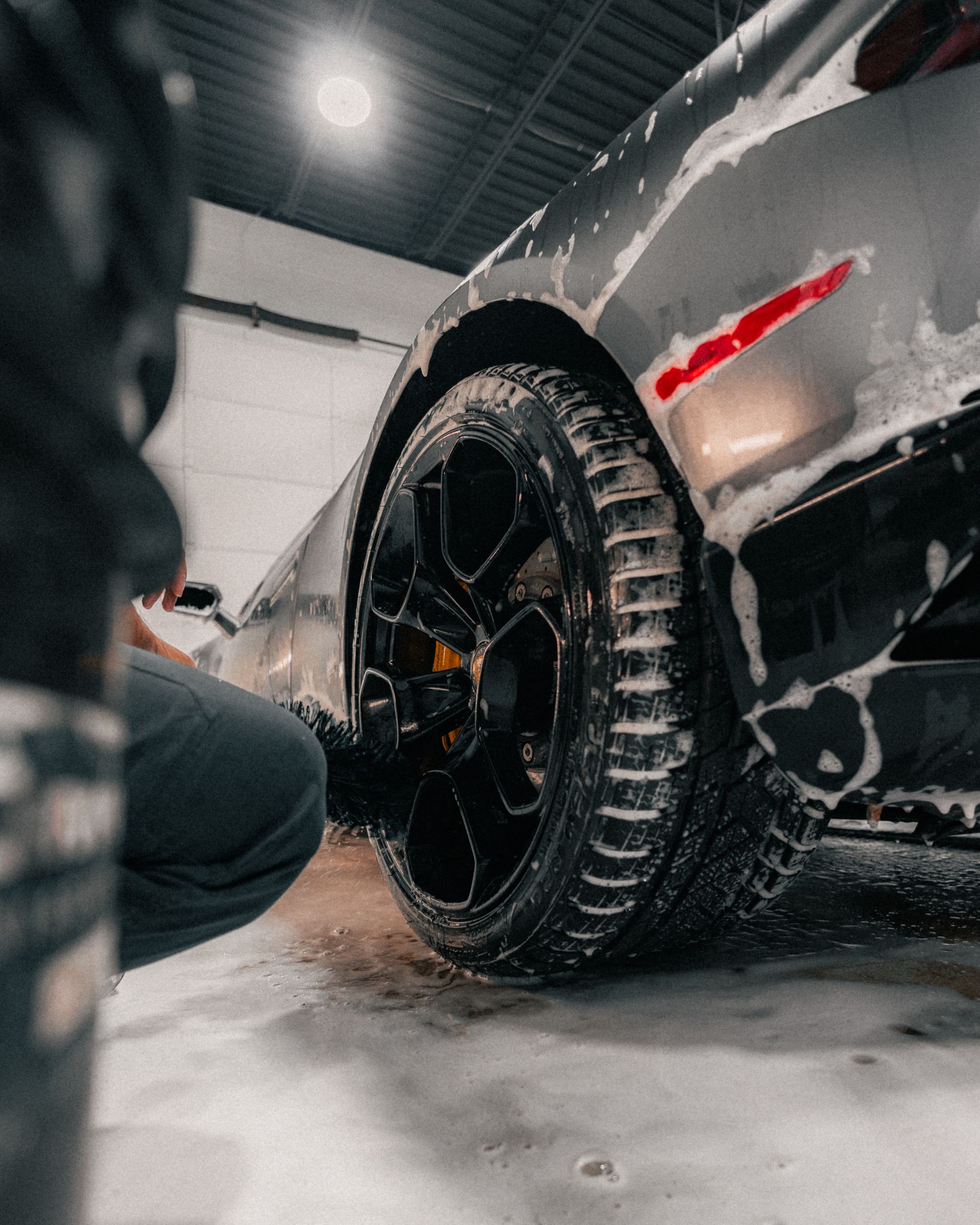Black sports car wheel in a garage, with a red side stripe and a person crouching nearby.